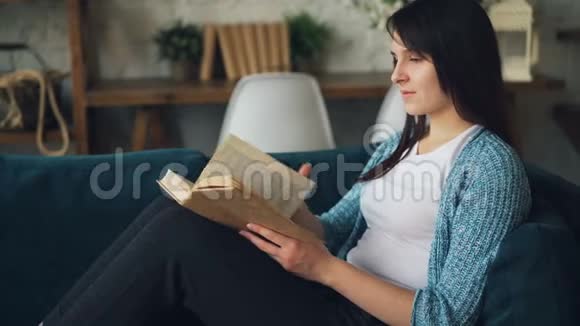 迷人的年轻女士正在看书微笑着坐在家里的沙发上独自放松有趣的文学视频的预览图