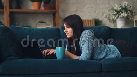 穿休闲服装的漂亮年轻女子正在笔记本电脑上打字在阁楼风格的工作室里躺在沙发上微笑视频的预览图