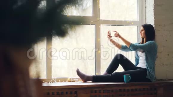 年轻的女士正在网上打视频电话坐在窗台上看着智能手机屏幕微笑着挥手视频的预览图