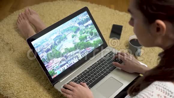 女孩用笔记本电脑在屏幕上观看大自然和度假视频而坐在家里的地毯上喝咖啡视频的预览图