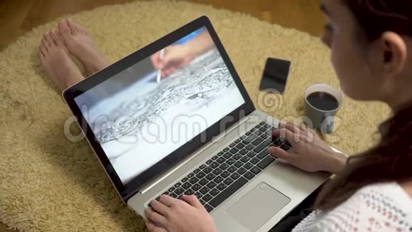 一位女士坐在地毯上的家里看着屏幕上有艺术素描视频的笔记本电脑视频的预览图