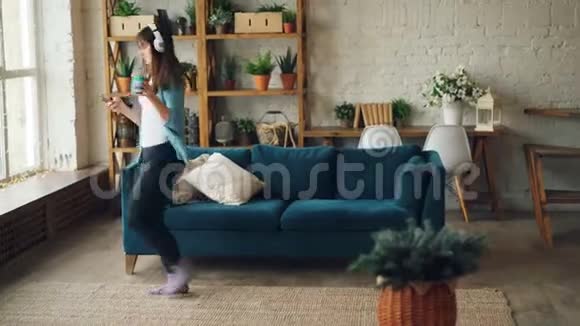 快乐的年轻女子拿着杯子和智能手机在家里跳舞戴着耳机听音乐享受着自由的时光视频的预览图