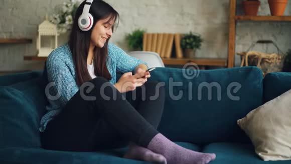 漂亮的女学生正在用耳机听音乐触摸智能手机屏幕在家坐着玩视频的预览图