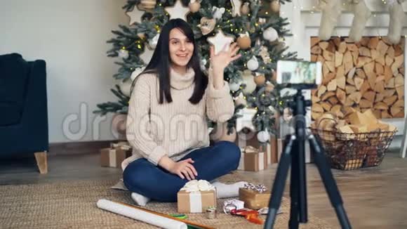 快乐的女人vlogger正在录制关于礼物包装的教程它坐在圣诞树附近拿着明亮的包装视频的预览图