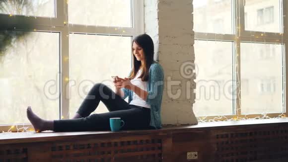 快乐的年轻女性正在使用智能手机坐在窗台上喝杯茶在家放松享受现代生活视频的预览图