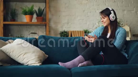 快乐的年轻女士正在通过耳机听音乐使用智能手机微笑和欢笑坐在沙发上视频的预览图