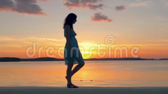 在夏日的傍晚美丽的女人穿着连衣裙沿着海滩漫步在夕阳下视频的预览图