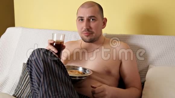 一个又胖又秃又懒的男人吃薯片喝可乐坐在沙发上没有外套一个卑鄙的人有害视频的预览图