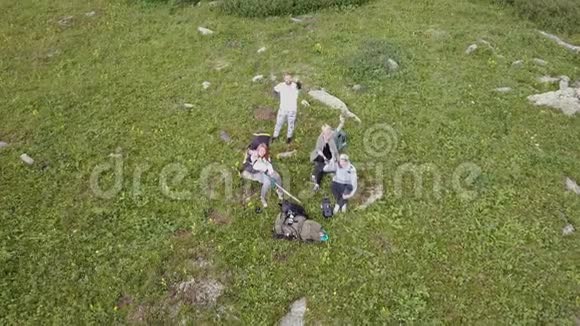 四名游客坐在高高的岩石山脚下的一个绿色的大空地上拍摄一段视频无人机飞离t视频的预览图