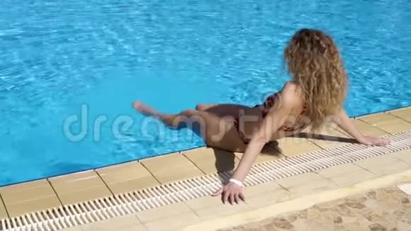 那个快乐的女孩坐在室外游泳池的边缘把脚伸进水里在泳池边休息一下度假村视频的预览图