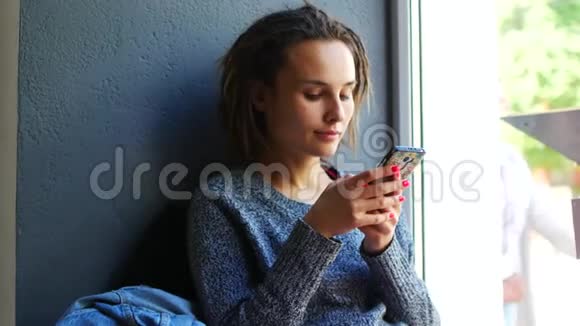年轻的白种人女孩在咖啡馆的窗户上享受移动智能手机视频的预览图