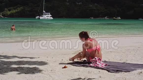 一个美丽的年轻女孩坐在海滩上用防晒霜涂抹她的身体视频的预览图
