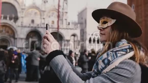 幸福的女人戴着时尚的帽子和白色的狂欢节面具在意大利威尼斯城市广场拍照视频的预览图
