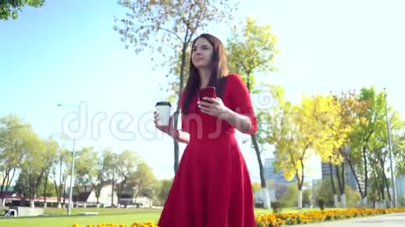 漂亮的商务女性穿着红色连衣裙在城市里散步喝咖啡用智能手机视频的预览图