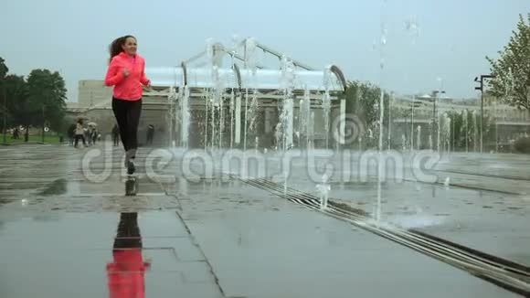 城市公园喷泉附近一个年轻的混血快乐女人在雨中奔跑的超慢动作镜头视频的预览图