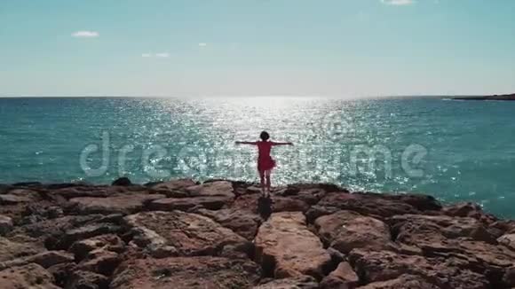 穿着红色连衣裙的女人剪影双臂分开在天空中站在珊瑚湾码头的岩石上看着大海和阳光的路反射视频的预览图