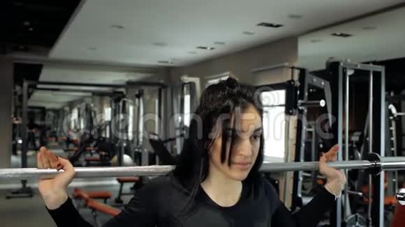 年轻的运动型黑发女孩准备用杠铃在蹲上做运动在健身房训练视频的预览图