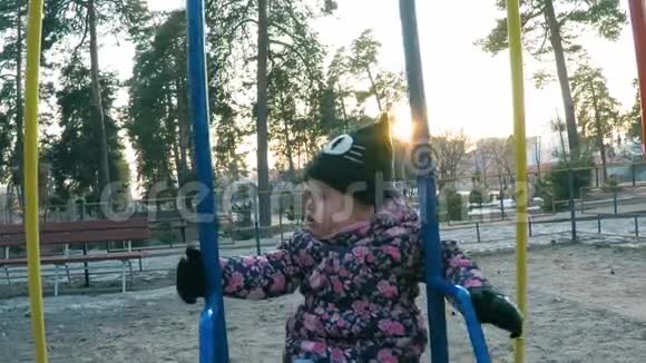 穿着五颜六色的粉红色夹克和黑色帽子的小女孩在公园的操场上骑着秋千早春的森林视频的预览图