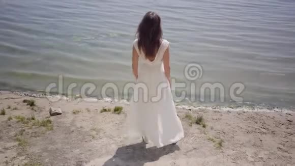 肖像迷人的年轻女孩留着长长的黑发穿着一件白色的夏季时装长裙站在河边视频的预览图