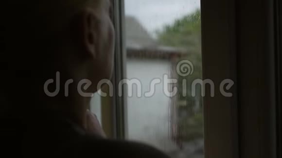 一个中年妇女站在窗前若有所思地透过玻璃看着雨视频的预览图