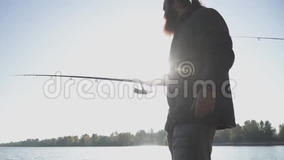 有胡子的渔夫正在阳光下的河岸上钓鱼人拿着鱼竿在河上钓鱼河流视频的预览图
