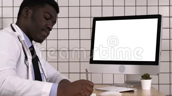 年轻的非洲医生穿着实验室外套坐在他的办公室里一边看电脑屏幕一边做一些笔记白色视频的预览图
