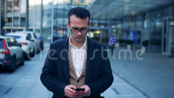 一位微笑的商人带着智能手机沿着现代城市街道行走的正面景色6K电影院的镜头视频的预览图