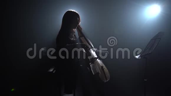 一个音乐女孩在一个黑暗的房间里排练大提琴演奏并揉进一个音乐摊剪影黑烟视频的预览图