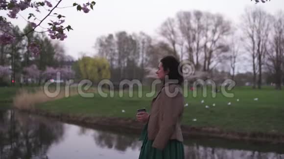 年轻女子走在公园里樱花盛开喝着纸杯里的咖啡做梦视频的预览图
