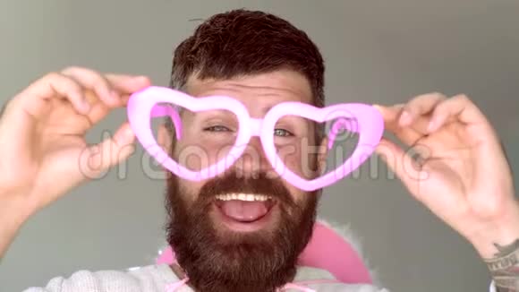 带着滑稽时髦的粉红色太阳镜和滑稽的粉红色帽子的疯子有趣的嬉皮士男人看着镜头在灰色视频的预览图