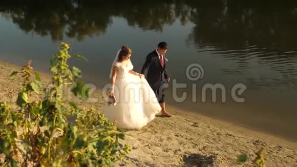 幸福的新娘穿着白色的衣服新郎穿着西装光着脚走在河岸边的沙子上爱的一对情侣要牵手视频的预览图