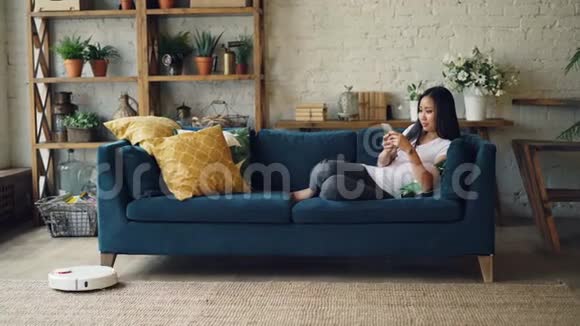 有魅力的亚洲女孩正享受着现代科技的乐趣她使用智能手机和机器人吸尘器坐在家里的沙发上视频的预览图