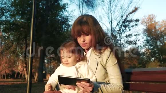 妈妈和孩子坐在公园里的长凳上在平板电脑里玩视频的预览图