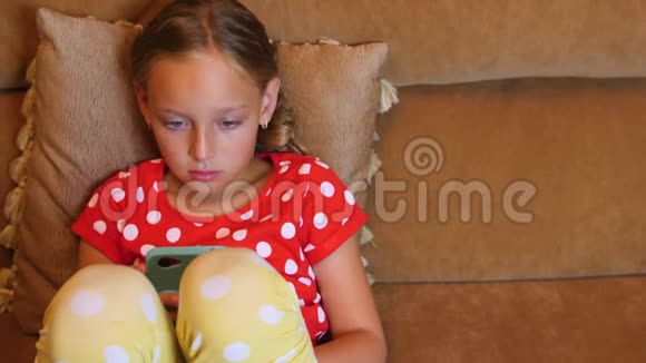小女孩坐在沙发上在房间里用手机在社交媒体上聊天视频的预览图