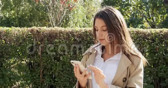 漂亮的黑发女孩呆在阳光下用她的智能手机看着相机微笑着阳光明媚的天气视频的预览图