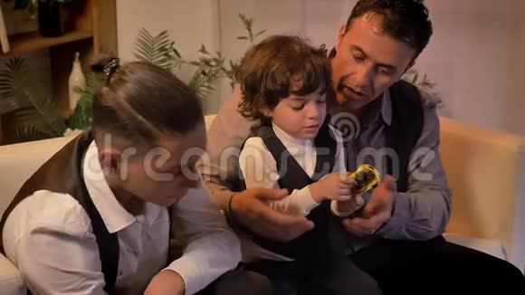 阿拉伯语的父亲和他十几岁的儿子坐在沙发上在客厅里和双胞胎卷发男孩玩耍视频的预览图