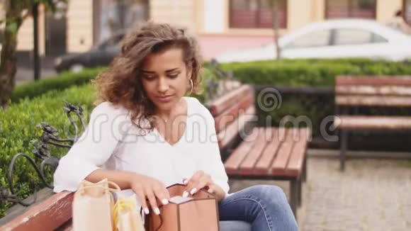 坐在长凳上购物后一位漂亮的女士正在看她买的东西视频的预览图