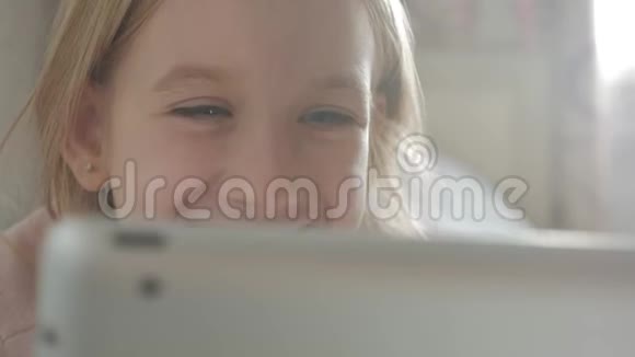 可爱的小女孩拿着数字平板电脑躺在床上微笑漂亮的女孩在平板电脑上玩教育视频的预览图