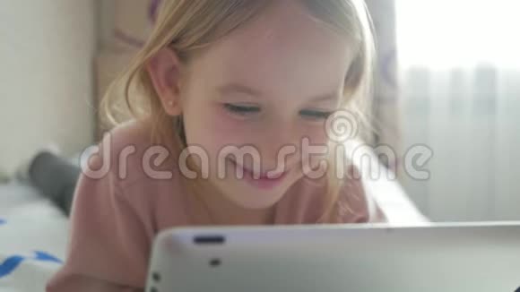 可爱的小女孩拿着数字平板电脑躺在床上微笑漂亮的女孩在平板电脑上玩教育视频的预览图
