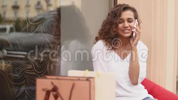 穿着时尚服装的优雅黑发女人坐在商场里通过电话交谈周围有许多五颜六色的购物袋阳光明媚视频的预览图