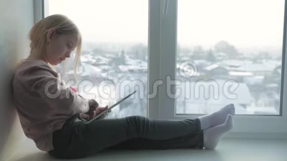 小女孩正坐在窗前的家中玩他们的技术平板电脑里面有游戏和娱乐应用程序视频的预览图