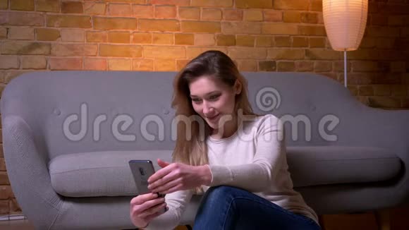 年轻迷人的白种人深色头发的女性用电话在室内舒适的地板上坐着的特写照片视频的预览图