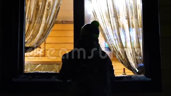 冬天的夜晚在靠近窗户的温暖明亮的房间里是一个年轻人敲着窗户在户外射击视频的预览图