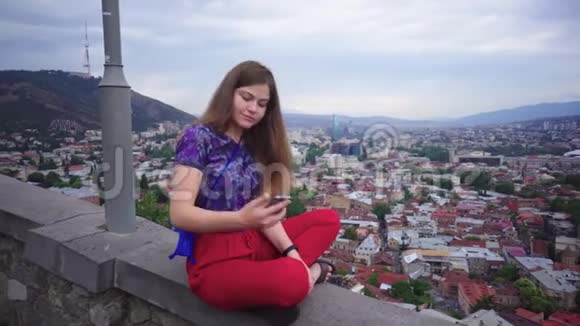 年轻的女游客在智能手机摄像头上拍摄带有城市景观美丽景色背景和微笑的自拍视频的预览图