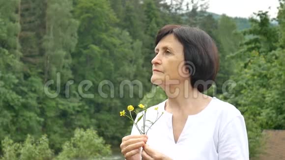 一位美丽的老年妇女在山林背景下的特写肖像手里拿着野花视频的预览图