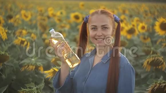 一个红头发的年轻女人拿着一瓶植物油站在田野里向日葵农业视频的预览图