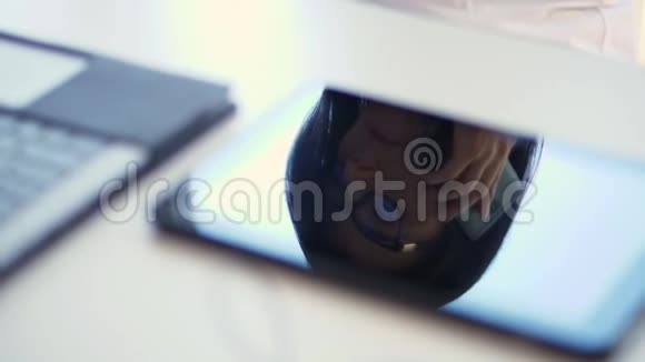 特写镜头在白色的桌面上在笔记本电脑旁边一个戴眼镜的商务女性的脸在平板电脑视频的预览图