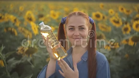 一个红头发的年轻女人拿着一瓶植物油站在田野里向日葵农业视频的预览图
