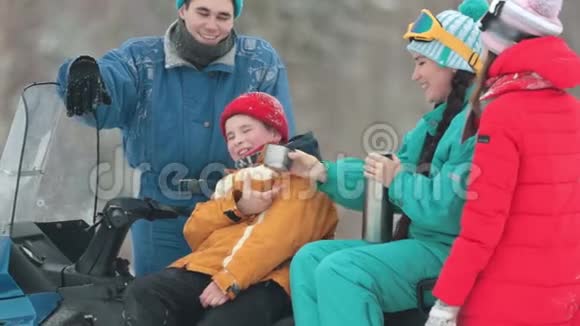 冬天一家人穿着五颜六色的衣服坐在雪地摩托上喝着泰莫斯的热饮一个小男孩视频的预览图
