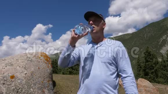 戴着帽子的人从山上和蓝天背景的瓶子里喝水视频的预览图